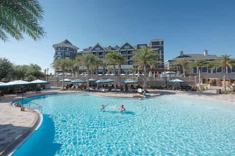 Resort courtyard pool with blue umbrellas, palm trees, and multi-story hotel building backdrop