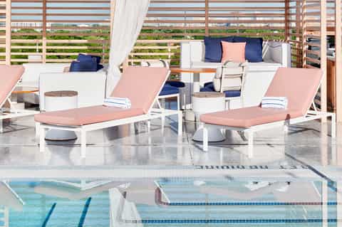 Poolside lounge area with pink cushioned loungers, white furniture, striped shade screen, and plush seating pods