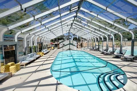 Indoor swimming pool under arched glass ceiling with white framework, lounge chairs, and bright natural light