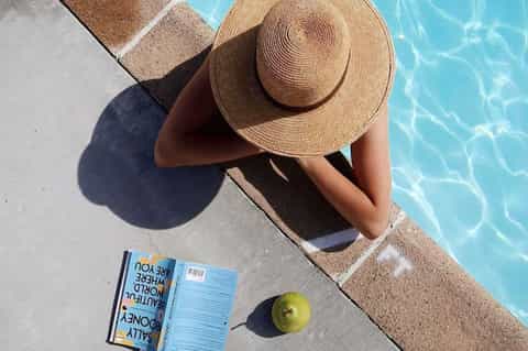 Person in straw hat by pool holding magazine and green apple