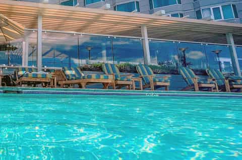 Resort pool with striped loungers, covered pergola, and ocean city view behind glass barrier