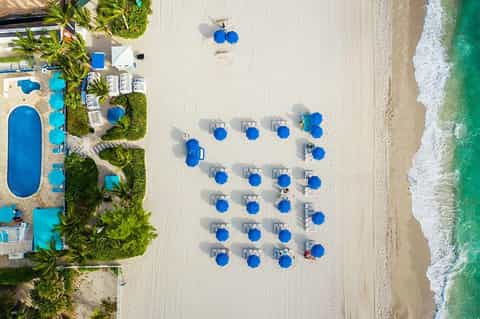 Aerial view of sandy beach with blue umbrellas, loungers, palm trees, and ocean waves
