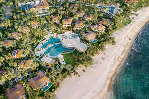 Aerial view of luxury beachfront resort with curved pool, palm-lined grounds, and pristine sandy beach