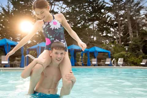 Girl on boy's shoulders in pool, girl wearing floral swimsuit, blue umbrellas in background
