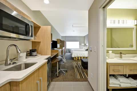 Modern hotel suite showing kitchenette, living area, bedroom with striped bedding, and bright bathroom with yellow accents