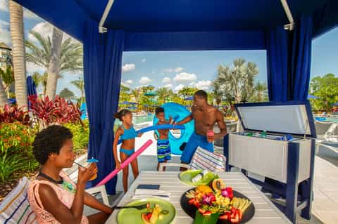Family enjoying poolside cabana with blue curtains, fresh fruit, tropical flowers, and water park view