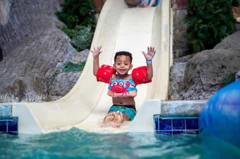 Young child sliding down waterslide into pool wearing red water wings and swim shirt