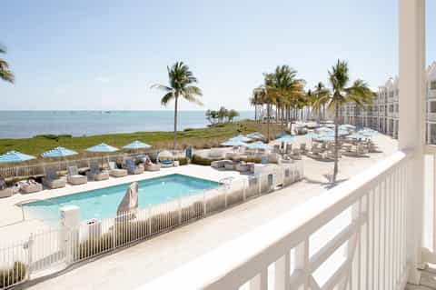 Beachfront resort pool surrounded by lounge chairs, umbrellas, palm trees, and ocean views with white railing