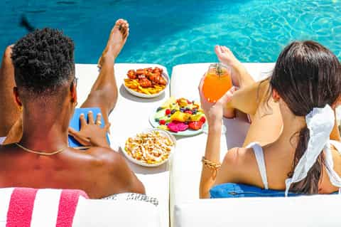 Guests relaxing poolside with colorful fruit platters and orange juice drinks by crystal blue water