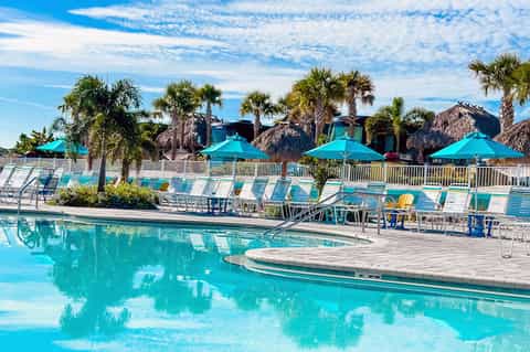 Resort pool with turquoise water, tiki umbrellas, loungers, and palm trees with thatched huts