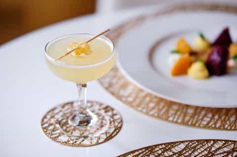 Elegant pale cocktail with gold garnish beside fine dining plate in upscale restaurant