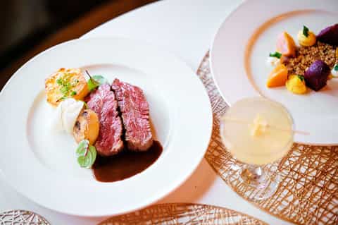 Gourmet plated dishes featuring seared meat and colorful vegetables with cocktail on textured placemat