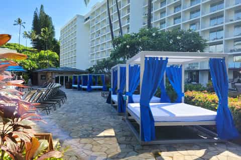 Resort pool deck with blue cabanas with curtains, white loungers, colorful flowers, and multi-story hotel building