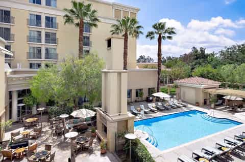Resort courtyard pool with lounge chairs, umbrellas, palm trees, and hotel building