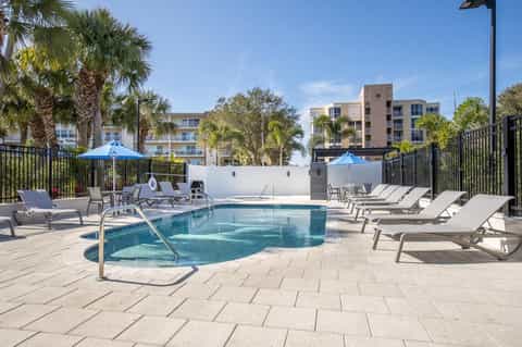Apartment complex rooftop pool with blue umbrellas, loungers, residential buildings, and palm trees against clear sky