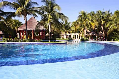 Resort swimming pool with thatched-roof cabanas, palm trees, and tropical landscape
