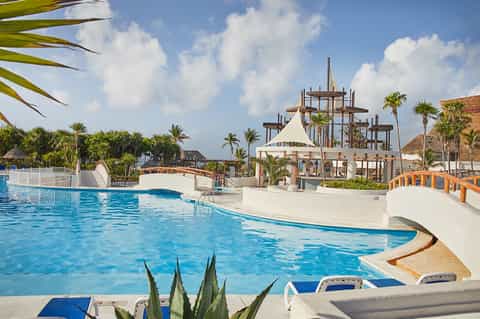 Large resort pool with waterslide tower, cabanas, and palm trees under blue sky