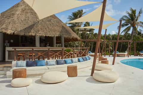 Tropical resort poolside lounge with cream seating, sail shade umbrellas, thatched bar, and palm trees
