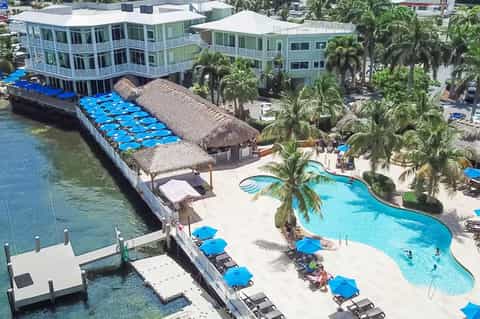 Aerial view of waterfront resort with pools, cabanas, palm trees, docked boat, and coastal buildings