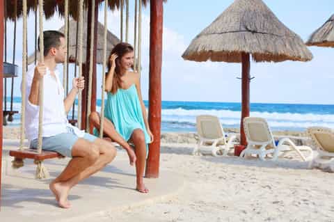 Couple on rope swing overlooking beach with thatched umbrella and lounge chairs in sand