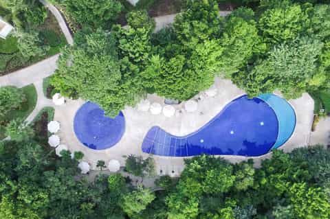 Aerial view of two curved blue resort pools nestled among lush green forest and white umbrellas