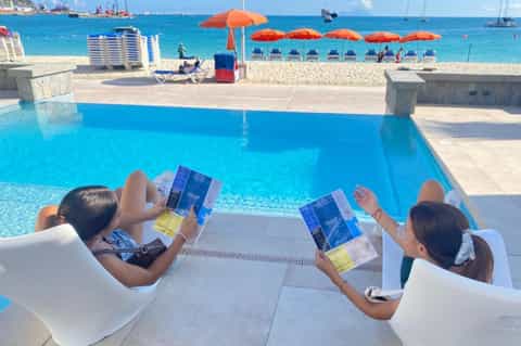 Resort beachfront pool with ocean view, lounge chairs, orange umbrellas, and sandy beach with umbrellas beyond