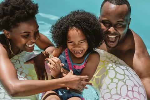 Family of three smiling and playing together in shallow turquoise ocean water under bright sunshine