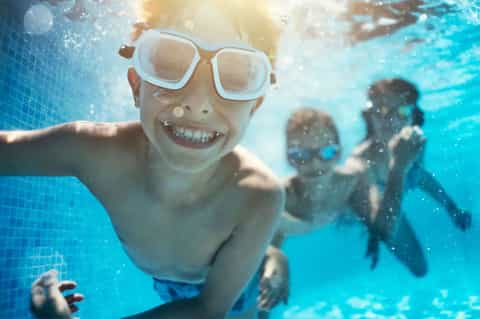 Child swimming underwater with goggles, smiling with other swimmers in background