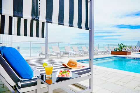 Beachfront cabana with striped awning, lounge chair, refreshments, and ocean view with pool in background
