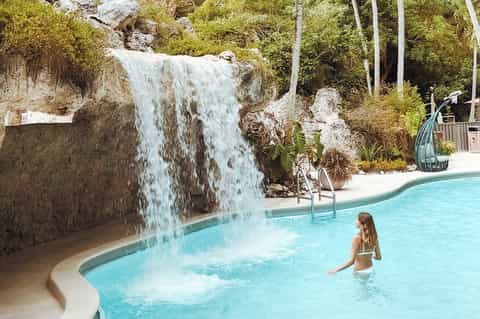 Woman in pool under cascading waterfall feature with tropical landscape and natural rock