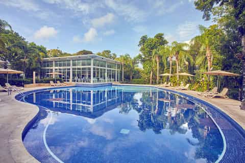 Resort pool with modern glass pavilion, palm trees, lounge chairs, and umbrellas on sunny day