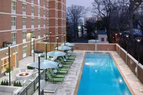 Urban rooftop pool at twilight with lounge chairs, fire pit, white umbrellas, and surrounding brick buildings