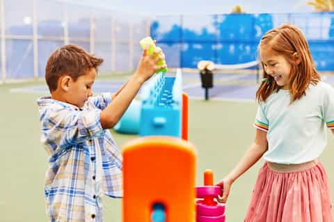 Two children playing with colorful toys on an outdoor tennis court with blue net