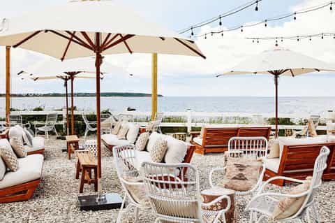 Beachfront lounge area with cream umbrellas, wooden daybeds, white chairs, and coastal view