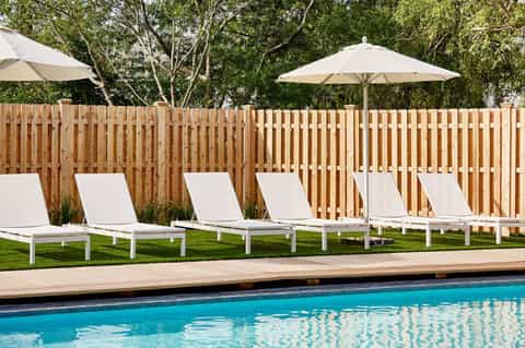Resort pool deck with white lounge chairs, umbrellas, wooden privacy fence, and manicured lawn