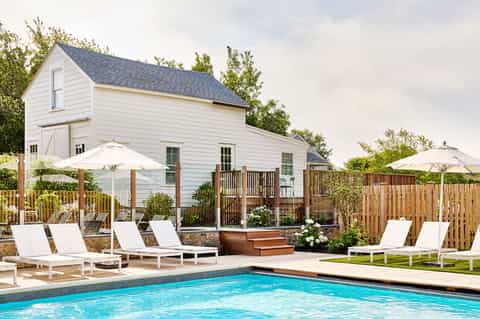 Resort pool with white loungers, umbrellas, and white farmhouse building surrounded by wooden fence