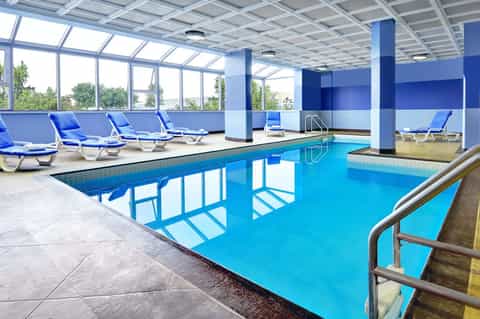 Indoor heated swimming pool with blue loungers and large windows showing natural light
