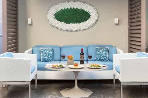 Contemporary outdoor lounge with blue sofa, white wicker chairs, artistic green wall art, and dining table