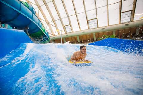 Indoor water park with surfing wave simulator, glass ceiling, and person boogie boarding