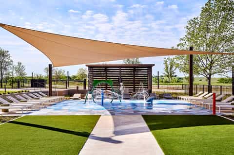 Outdoor splash pad with blue water features, tan shade sail, wooden shelter, and landscaped grounds