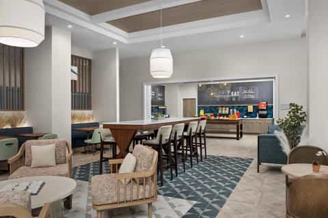 Modern hotel lounge and bar area with wooden furnishings, comfortable seating, and open kitchen setup