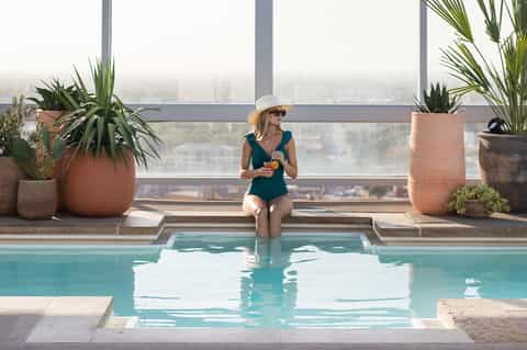 Woman in swimsuit sitting at edge of high-rise rooftop pool with city skyline view behind her