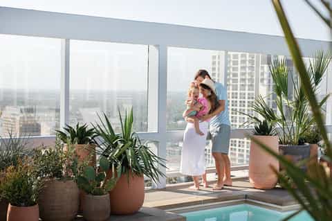 Family enjoying high-rise balcony view with potted plants overlooking cityscape and distant pool