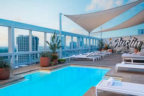 Rooftop pool overlooking city skyline with sail shade, loungers, and urban landscape view