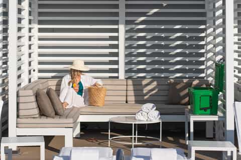 Woman sitting on modern white lounge seat on resort balcony with striped privacy screen and rolled towels