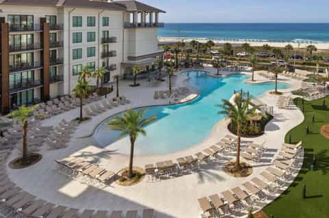 Large curved lagoon-style pool with beach beyond, palm trees, loungers, and modern beachfront resort buildings