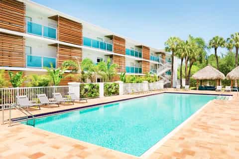 Resort pool with turquoise water, white lounge chairs, modern multi-story building with wood slats, and palm trees