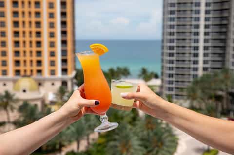 Two tropical cocktails held aloft with ocean and high-rise hotels in background