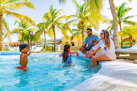 Family enjoying resort pool with children playing in water under palm trees and yellow buildings