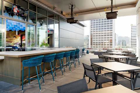 Rooftop bar and lounge with blue bar seating, tables, city skyline views, and floor-to-ceiling windows